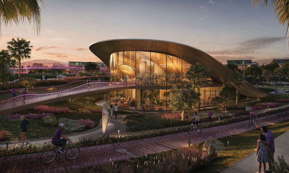 Evening view of a curved glass pavilion, a footbridge and cyclists on a planted path at Athlon in Dubailand.
